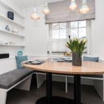 Rundtisch mit Sitzbank, Bücherregal und Fenster mit Blumen.