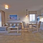 Wohnbereich mit Sofa, Esstisch und Küchenzeile. Holzboden und große Fenster.