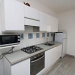 Küche mit weißen Schränken, Herd, Mikrowelle und Kühlschrank. Durchgang zur Terrasse.