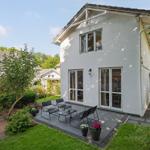 Weiße Villa mit Terrasse und Garten. Glastüren führen ins Haus. Möbel und Pflanzen sind auf der Terrasse.