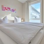 Ein Bett mit weißen Kissen und Bettwäsche. An der Wand hängt ein buntes Bild. Ein Fenster mit weißen Vorhängen.