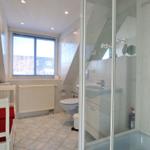 Badezimmer mit Dusche, Toilette und Fenster. Ein weißer Kinderstuhl mit rotem Kissen steht daneben.