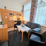 Wohnbereich mit Sofa, Tisch, Küchenzeile und Fenster mit Vorhängen.