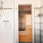 Badezimmer mit Dusche, Waschbecken und Glasregal. Durchgang zu einem Raum mit Kleiderständer.