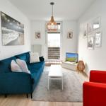 Wohnzimmer mit blauem Sofa, rotem Sessel und Fernseher. Helle Wände und Holzboden.