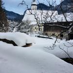 Snow-covered church with tower in winter, surrounded by mountains and bare branches.