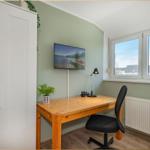 Ein Arbeitszimmer mit Schreibtisch, Stuhl und Fernseher an der Wand. Fenster mit Blick nach draußen.