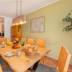 Essbereich mit Holztisch, Stühlen und Obst. Küche im Hintergrund. Wandbild und Zimmerpflanze.