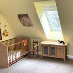Kinderzimmer mit Babybett, Stuhl und Schrank unter Dachschräge mit Fenster.