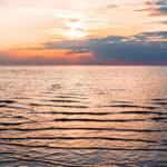 Sonnenuntergang über ruhigem Wasser mit sanften Wellen und dunklen Pfählen im Hintergrund.
