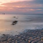 Sonnenuntergang über ruhigem Wasser mit Sandstrand und entfernten Windrädern.