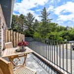 Terrasse mit Holztisch, Stühlen und Blumen. Blick auf grüne Bäume und einen gepflasterten Weg.