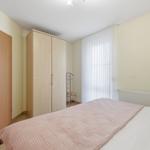 Schlafzimmer mit Bett, Kleiderschrank und Fenster mit weißen Vorhängen.