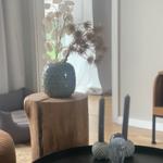 Ein grauer Vase mit Trockenpflanzen steht auf einem Holzblock. Vorherige Tisch mit Kerzen und Glasobjekten.