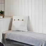 Badezimmerbank mit Kissen und Decke, Wand mit vertikalen Paneelen, Pflanze und Kerze auf Oberfläche.