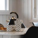 Tisch mit Kanne, Tassen und Keksen vor Fenstern.