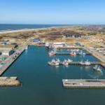 Hafen mit Booten, Industriegebäude und Windkraftanlage am Strand.