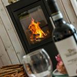 Kamin mit brennendem Feuer, Weinflasche und Glas auf Holzwand im Vordergrund.