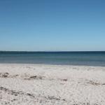 Weißer Sandstrand mit Blick auf Meer und Brücke im Hintergrund.