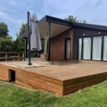 Moderne Villa mit Holzterrasse und Sonnenschirm. Grün umgeben.