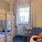 Zimmer mit Doppelstockbett, Schreibtisch und Stühlen. Fenster mit Vorhängen. Wandbild.