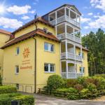 Gelbes Mehrfamilienhaus mit weißen Balkonen und rotem Dach. Umgeben von Bäumen und Grünflächen.