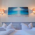 Ein Bett mit weißen Kissen und Handtüchern. Über dem Kopfteil hängt ein Strandbild mit zwei Wandlampen.