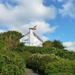 Weißes Haus mit Flagge auf Hügel, umgeben von grüner Vegetation und blauem Himmel.