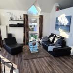 Wohnzimmer mit schwarzen Ledermöbeln, Glas-Tisch und Holzboden. Fenster mit Vorhängen.