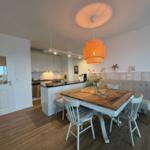 Essbereich mit Holztisch, weißen Stühlen und orangefarbener Hängelampe. Küche im Hintergrund.
