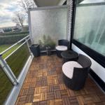 Terrasse mit zwei Stühlen und Tisch, Holzbelag, Glasgeländer und Sicht auf Grünfläche.