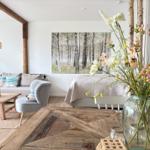 Wohnraum mit Sofa, Stuhl und Holztisch. Wandbild zeigt Birkenwald. Blumen im Glasvasen.