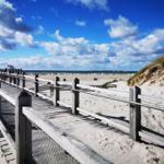 Holzbrücke führt zum Strand. Himmel blau mit weißen Wolken. Dünen und Sandstrand im Hintergrund.