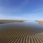 Sandflächen mit Wellenmuster und Wasserpfad unter blauem Himmel