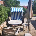 Terrasse mit Strandkorb, Tisch und Stühlen. Hintergrund mit Bäumen und Steinmauer.