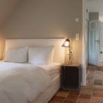 Schlafzimmer mit Bett, Nachttisch und Stehlampe. Blick in den Flur mit Terrakottaboden.