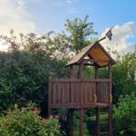 Holzspielhaus mit Flagge im Garten, umgeben von Bäumen und Rosen.