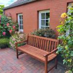 Holzbank auf gepflasterter Terrasse vor rotem Backsteinhaus mit Rosenbeete und Fenstern.