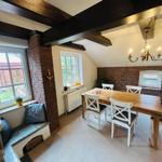 Esszimmer mit Holztisch, weißen Stühlen und Backsteinwand. Deckenbalken und Kronleuchter sichtbar.