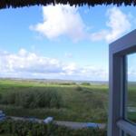 Blick aus dem Fenster auf grünes Feld und Meer unter blauem Himmel mit weißen Wolken.