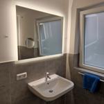 Badezimmer mit Waschbecken, beleuchtetem Spiegel und blauen Handtüchern an der Wand.