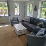 Wohnzimmer mit Sofa, zwei Sesseln und Holztisch. Fenster bieten Blick auf Garten.