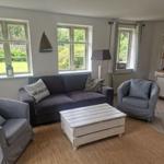 Wohnzimmer mit Sofa, zwei Sesseln und Holztisch. Fenster zeigen Garten. Wanduhr und Schrank sichtbar.