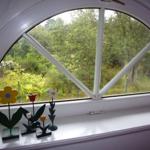 Blick durch ein rundes Fenster auf einen grünen Garten mit Blumenfiguren auf dem Fensterbrett.