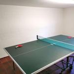 Tischtennisplatte mit Netz und Schlägern in einem Raum mit weißen Wänden und gefliestem Boden.