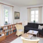 Wohnzimmer mit Sofa, Bücherregal, Fernseher und zwei Fenstern.