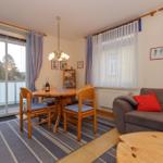 Wohn- und Essbereich mit Sofa, Tisch und Stühlen, Balkon und Fenster mit Vorhängen.