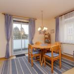Esszimmer mit Holztisch, Stühlen und Balkontür. Sofa und Bücherregal sind sichtbar.