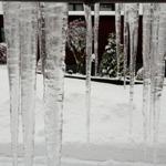 Eisstürze hängen von einem Dach, Schnee bedeckt den Boden und Bäume im Hintergrund.