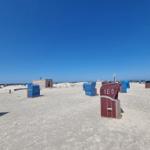 Strand mit bunten Strandkörben und kleinem Holzhaus unter blauem Himmel.